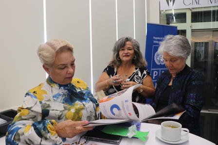 Three women seated at a table, two reading magazines and one gesturing while smiling.