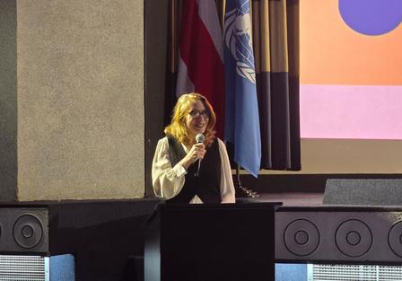 A speaker with red hair stands at a podium, addressing an audience with flags behind.