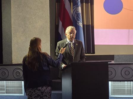 A man in a suit stands at a podium while a woman gestures during a discussion.