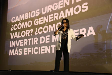 A speaker in a formal outfit stands on stage, engaging with the audience against a backdrop of text.