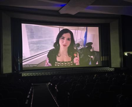 A woman speaks on a large screen in a nearly empty theater.