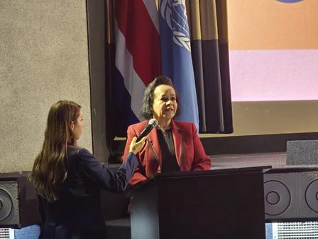 A woman in a red blazer speaks at a podium while another woman holds a microphone.