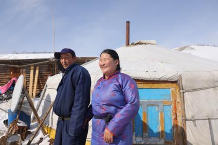 Two people stand beside a traditional yurt in a snowy landscape, smiling and dressed warmly.