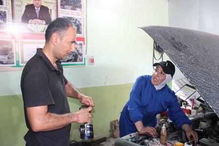 A mechanic in blue overalls works on a car engine while a man observes.