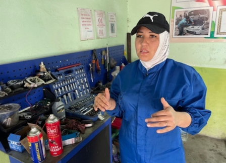 A woman in a blue coverall and hat gestures near a tool station in a workshop.