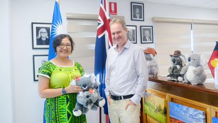 Two people pose with koala plush toys, flags in the background, smiling in a bright room.