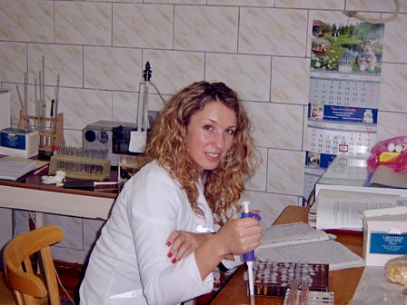 A woman with curly hair in a lab coat holds a pipette at a cluttered laboratory table.