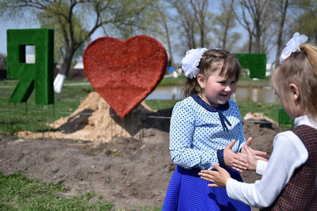Two girls play and laugh in front of a large heart sign on a sunny day.
