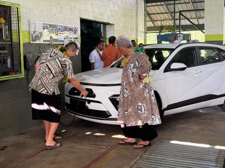 Two women charging a white electric vehicle in a service station, with others in the background.