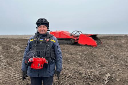 A smiling man in a tactical vest stands in a barren field, with machinery in the background.