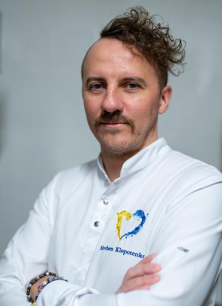 A man with curly hair and a mustache stands confidently, wearing a white shirt with a logo.