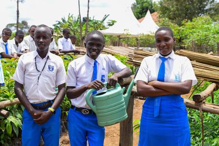 Three students in uniforms stand in a garden, one holding a watering can, smiling.