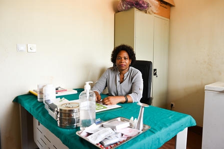 A woman with curly hair sits at a desk covered with medical supplies in a clinic.