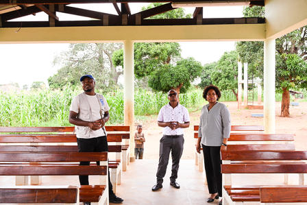 Three people stand in an empty indoor space with wooden benches, greenery visible outside.