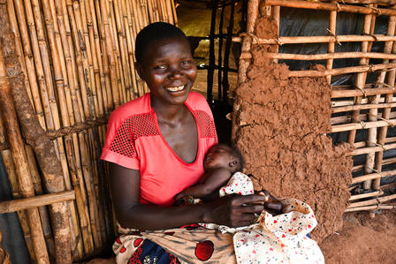 Smiling woman in a red shirt sitting against a rustic wall, holding a baby.