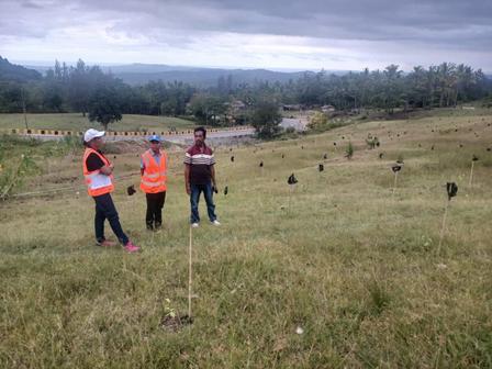 Three individuals in safety vests stand in a grassy field with young trees planted around.
