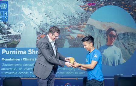 A man in a suit hands a trophy to a smiling individual in a blue shirt against a backdrop of mountains.