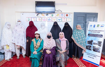 A diverse group of women in traditional attire poses for a photo in a classroom setting.