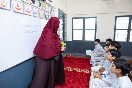 A teacher in a maroon dress interacts with attentive students in a classroom setting.