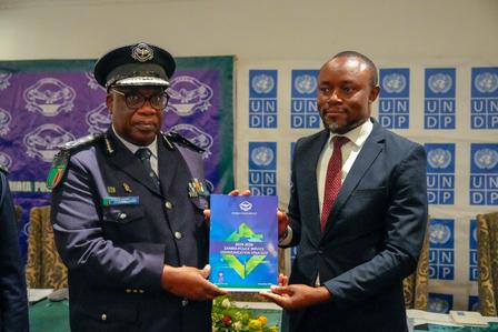 Two men in formal attire exchange a document, smiling, with a UNDP backdrop.
