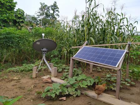 A solar panel and satellite dish set up in a green field with maize plants.