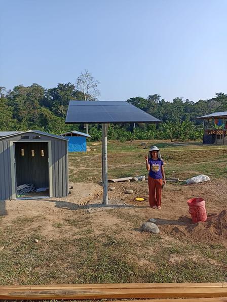 A young woman stands next to a solar panel setup and a small shed in a rural area.