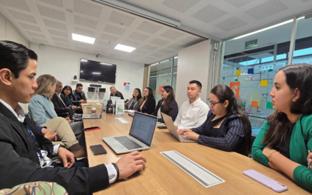 A group of diverse individuals seated around a conference table, engaged in discussion with laptops.