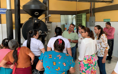 Group of people engaged in discussion around a coffee roasting machine in a bright room.