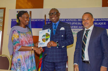 A woman hands over a document to a man in a suit, while another man in a suit observes.