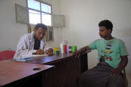 A doctor in a white coat writes notes while a patient sits across from him in a clinic.