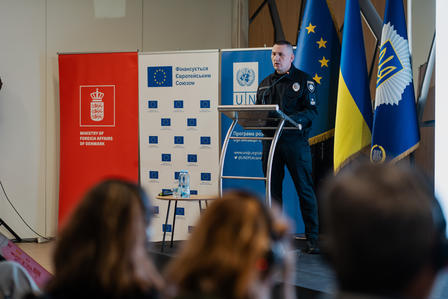 A police officer speaking at a podium with flags and banners in a conference setting.