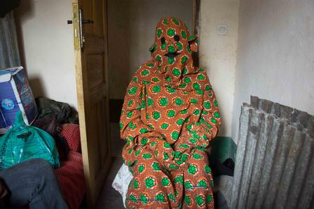 A person in vibrant orange and green patterned clothing sits against a wall in a dim room.