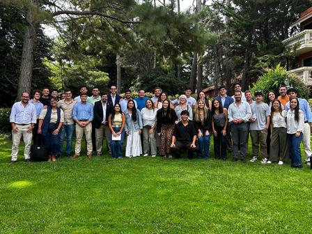 A large group of people gathers on a green lawn surrounded by trees.