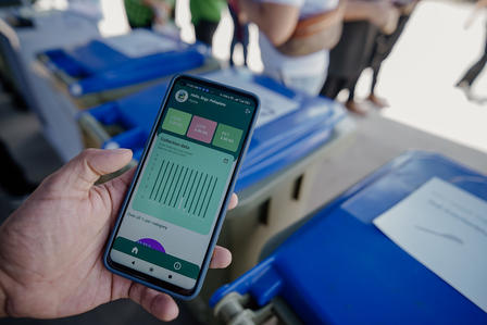 A hand holding a smartphone displaying a waste management app, with blue bins in the background.
