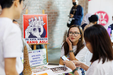 A woman with glasses listens intently at a table with signage in a lively gathering.