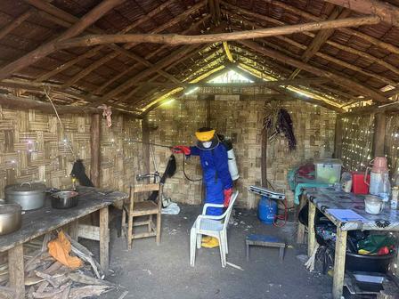 A person in protective gear sprays disinfectant inside a simple bamboo structure.
