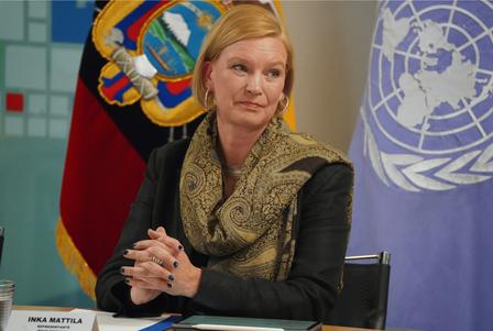 A woman with blonde hair, wearing a patterned scarf, sits at a table with flags in the background.