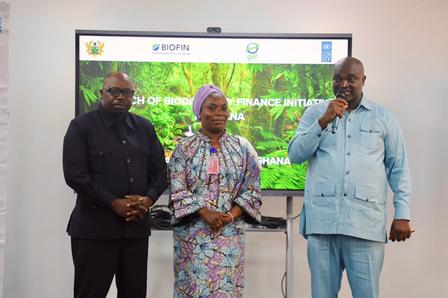 Three professionals stand together in front of a screen displaying greenery, one speaking.