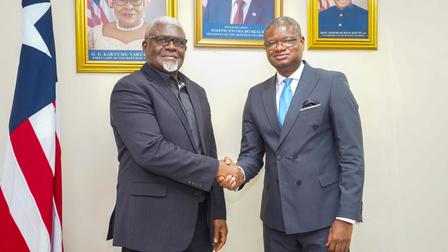 Two men in business suits shaking hands, with a flag and portraits in the background.