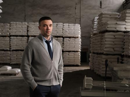 A man in a grey sweater and tie stands in a warehouse surrounded by stacked bags.