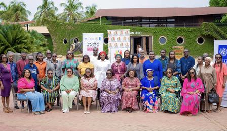 Group photo of diverse individuals, smiling, outdoors with greenery and a building in the background.