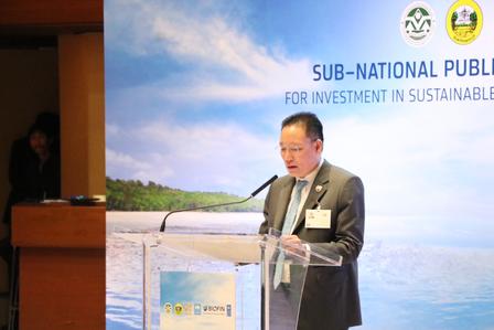 A man speaking at a podium during a conference with a backdrop of blue sky and greenery.
