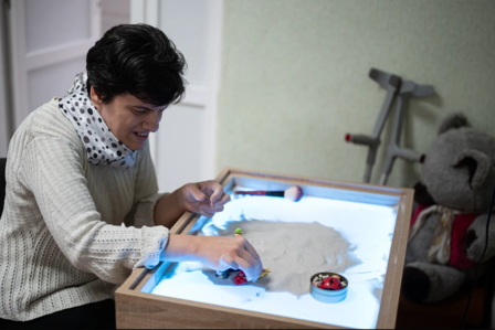 Woman playing with sand on a light table, surrounded by toys and a crutch.
