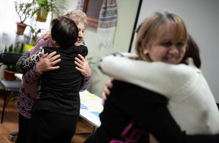Four people embrace warmly in a cozy indoor setting with plants in the background.