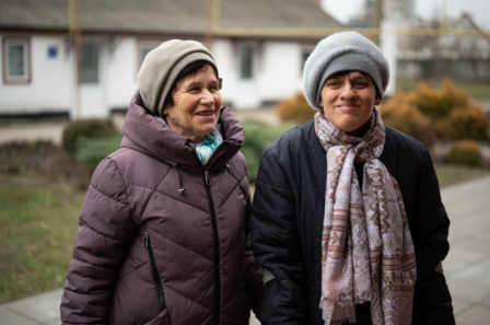 Two women smiling outdoors, wearing winter coats and hats, with buildings and greenery in the background.