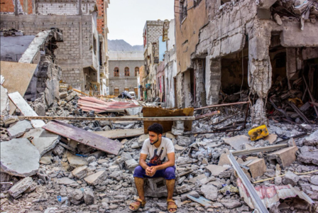 A man sits on rubble amid the wreckage of collapsed buildings.