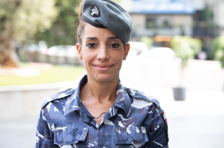 Smiling woman in a military uniform with a beret, standing outdoors.