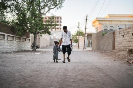 A man and a child walking hand in hand down a dusty street at sunset.