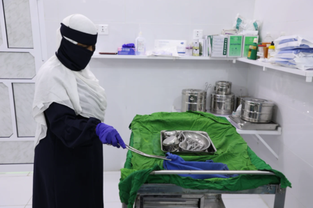 A person in a hijab and gloves stands by a table with a green cloth in a clinical setting.