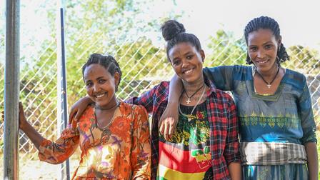 Three young women smiling together outdoors, standing by a fence.